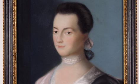 A framed portrait of a young woman with dark hair. She wears a blue dress with lace around the neckline, a pink wrap, a pearl choker necklace, and a pink ribbon in her hair.